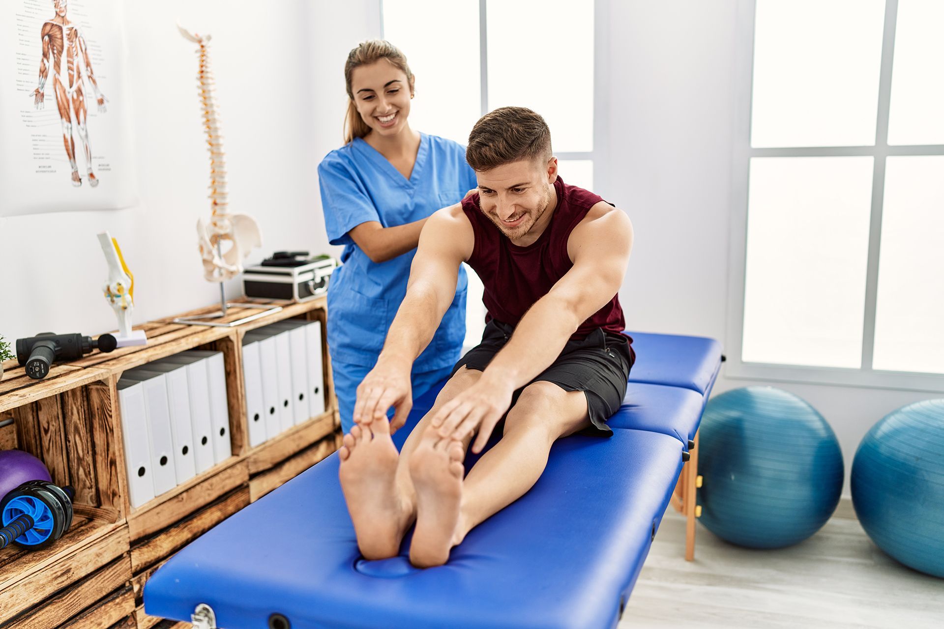 A man is sitting on a blue table stretching his legs while a nurse looks on.