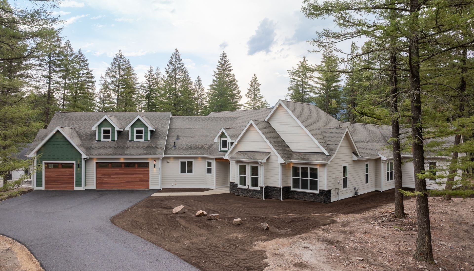 Large gray house with green-trimmed dormers, red garage doors, and a gravel driveway in a pine forest setting