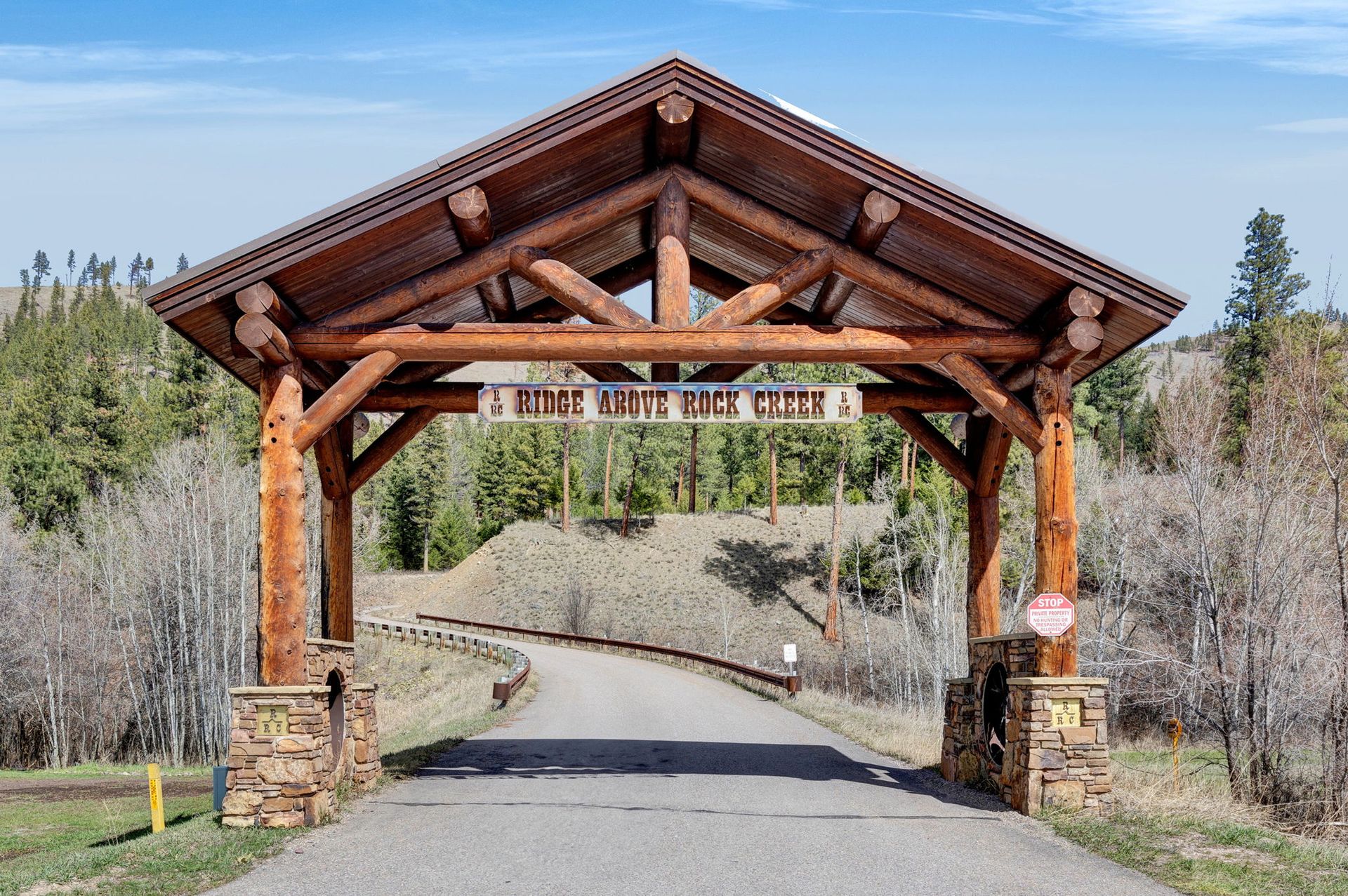 Wooden entrance arch over a paved road in a wooded area with a banner hanging across it