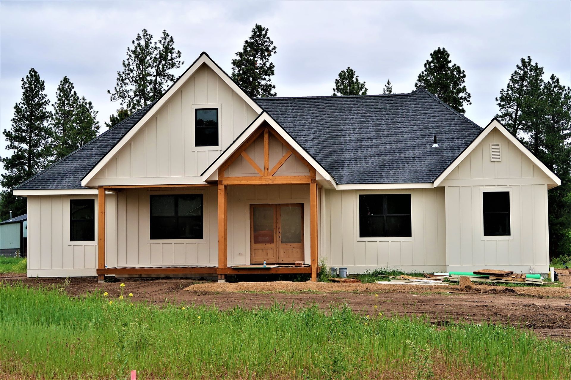 New beige house with dark roof and wooden front porch, surrounded by grass and trees