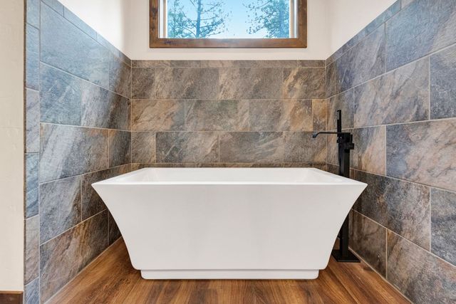 Modern freestanding bathtub in a tiled bathroom with wood floor and a small window above.