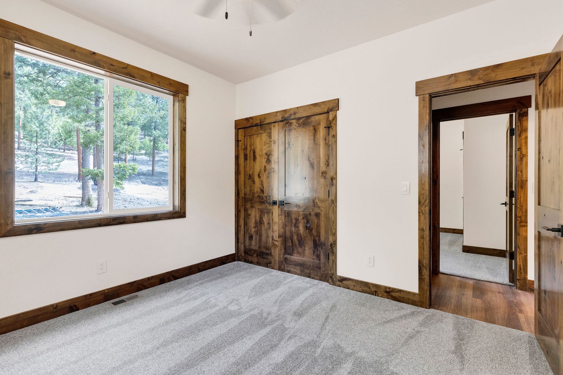 Empty room with gray carpet, white walls, wood trim, rustic door, and large window with trees outside