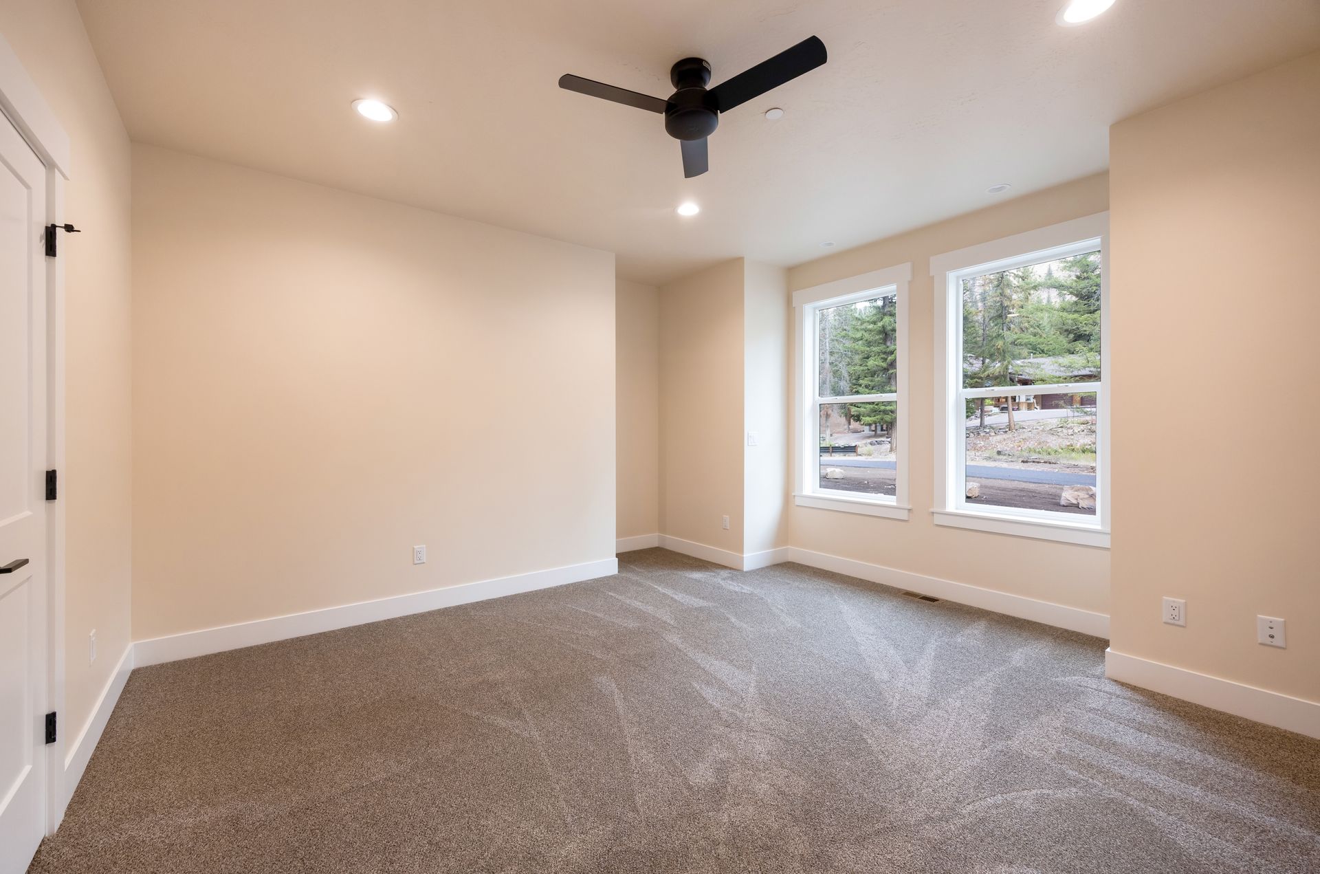 Empty beige bedroom with carpet, black ceiling fan, and two windows overlooking greenery.