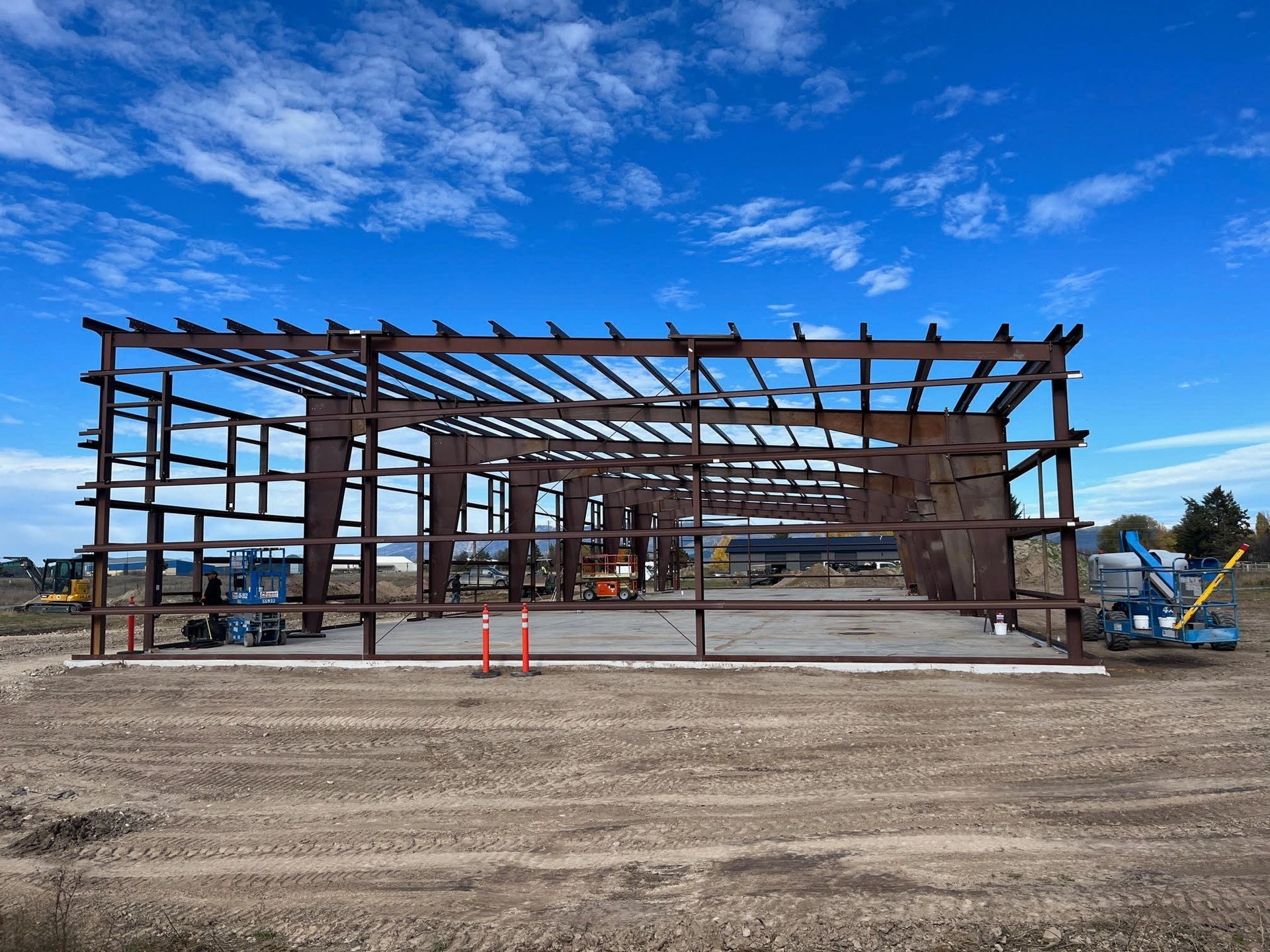 Steel frame of a building under construction on a dirt lot beneath a blue sky.