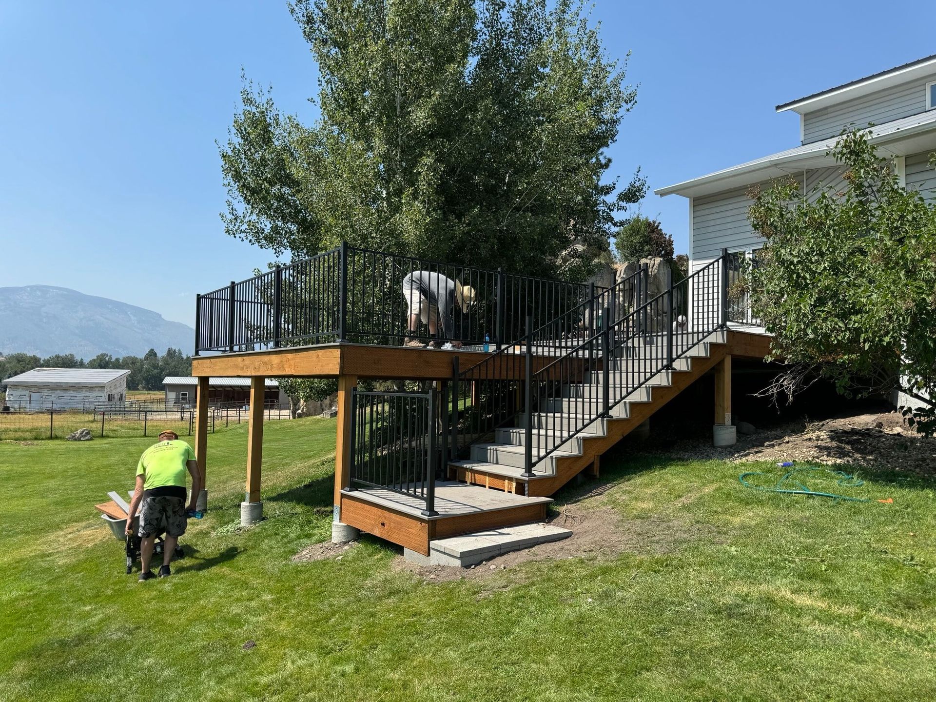 Deck with stairs beside a house, with a worker kneeling on the lawn and a large tree in the yard