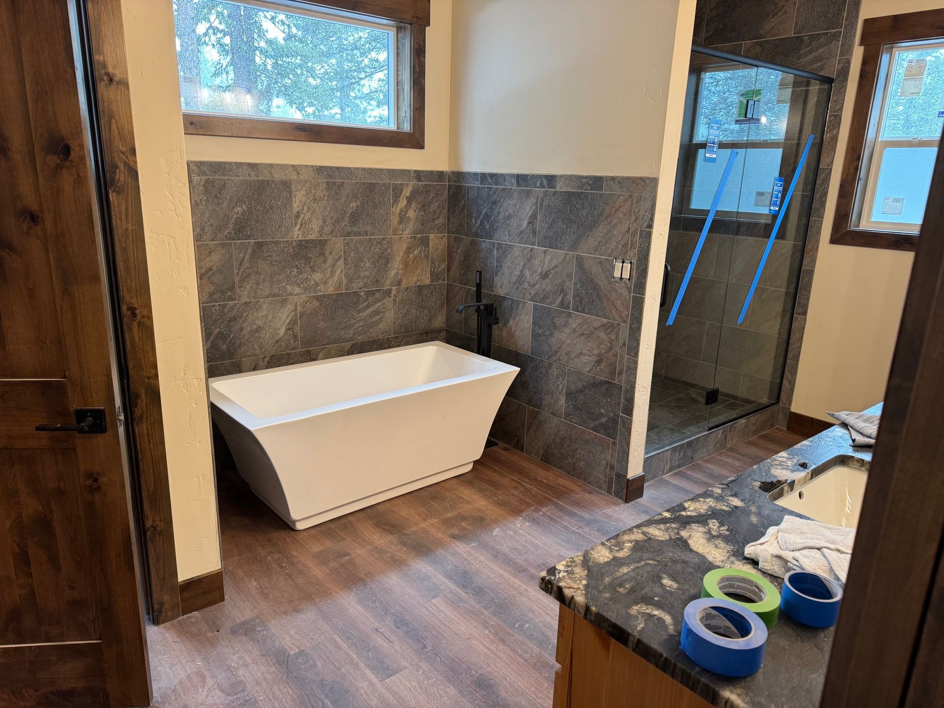 Modern bathroom with freestanding white tub, dark tile walls, wood floor, and glass shower.