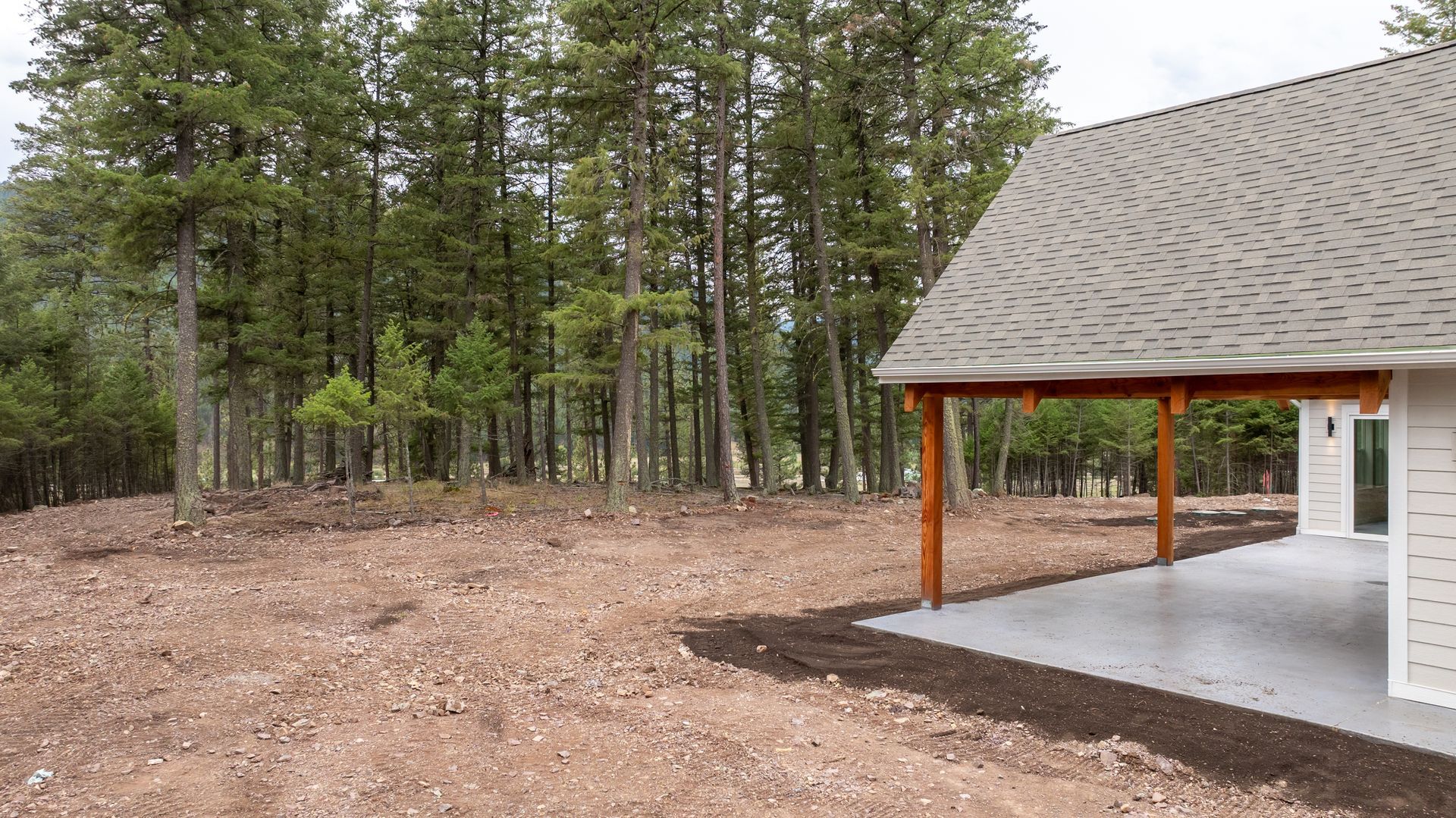 Forest clearing beside a covered pavilion with wooden posts and a gray shingled roof