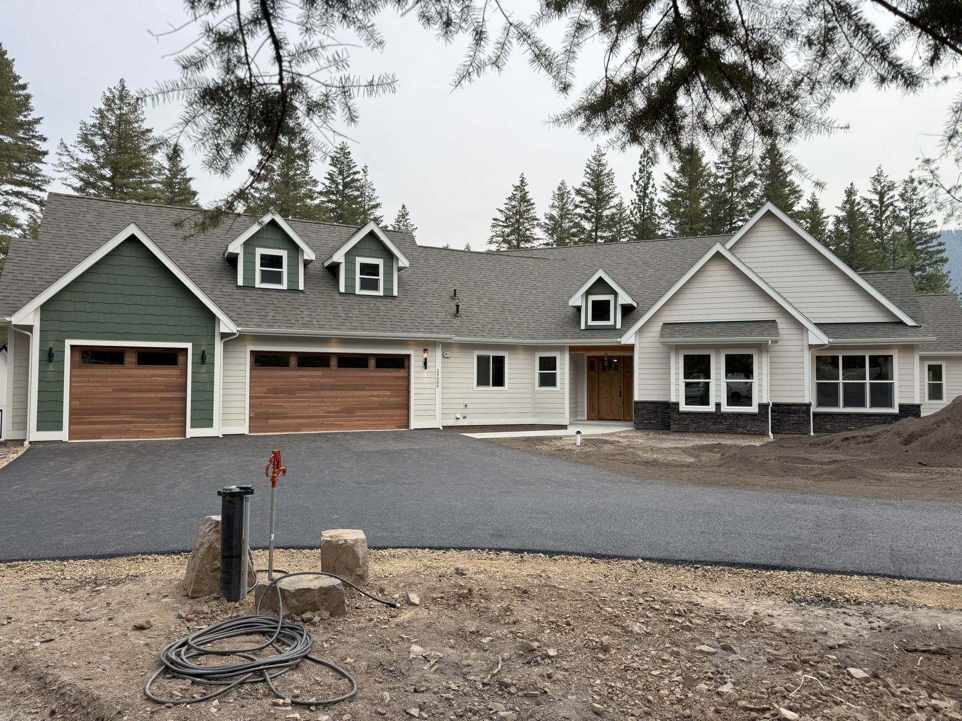Large gray house with green trim and three garages, viewed from a gravel driveway under overcast skies