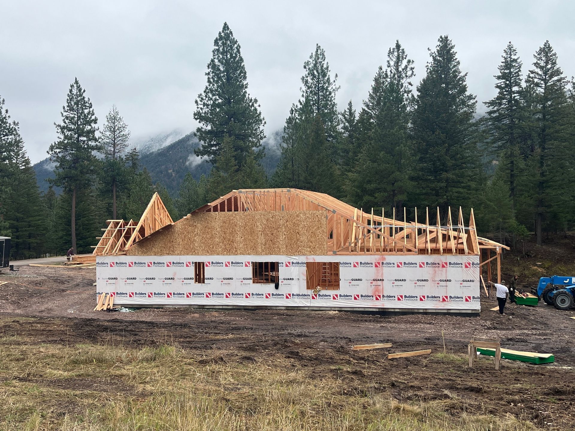 House under construction with exposed wooden framing and white wrap in a forested mountain setting