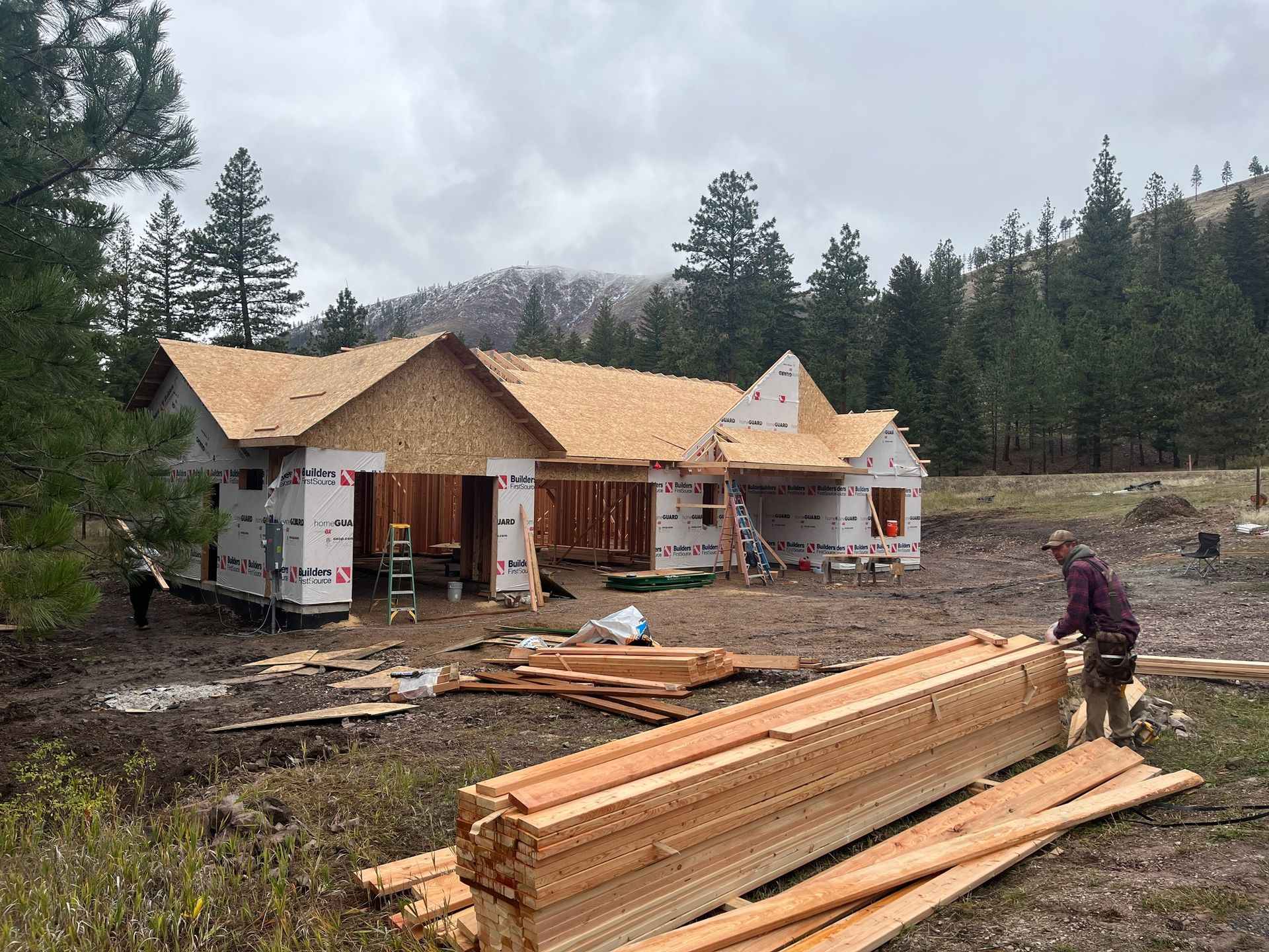 House under construction with workers and lumber stack in a muddy forest clearing