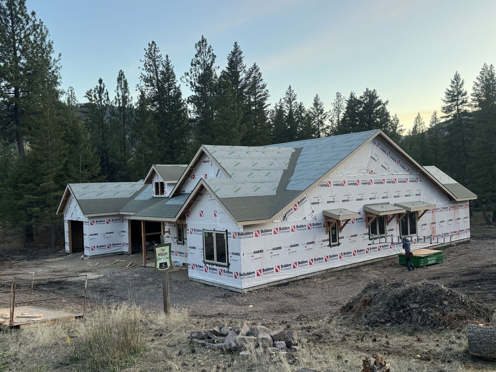 Partially built house wrapped in house wrap, surrounded by dirt and pine trees at dusk