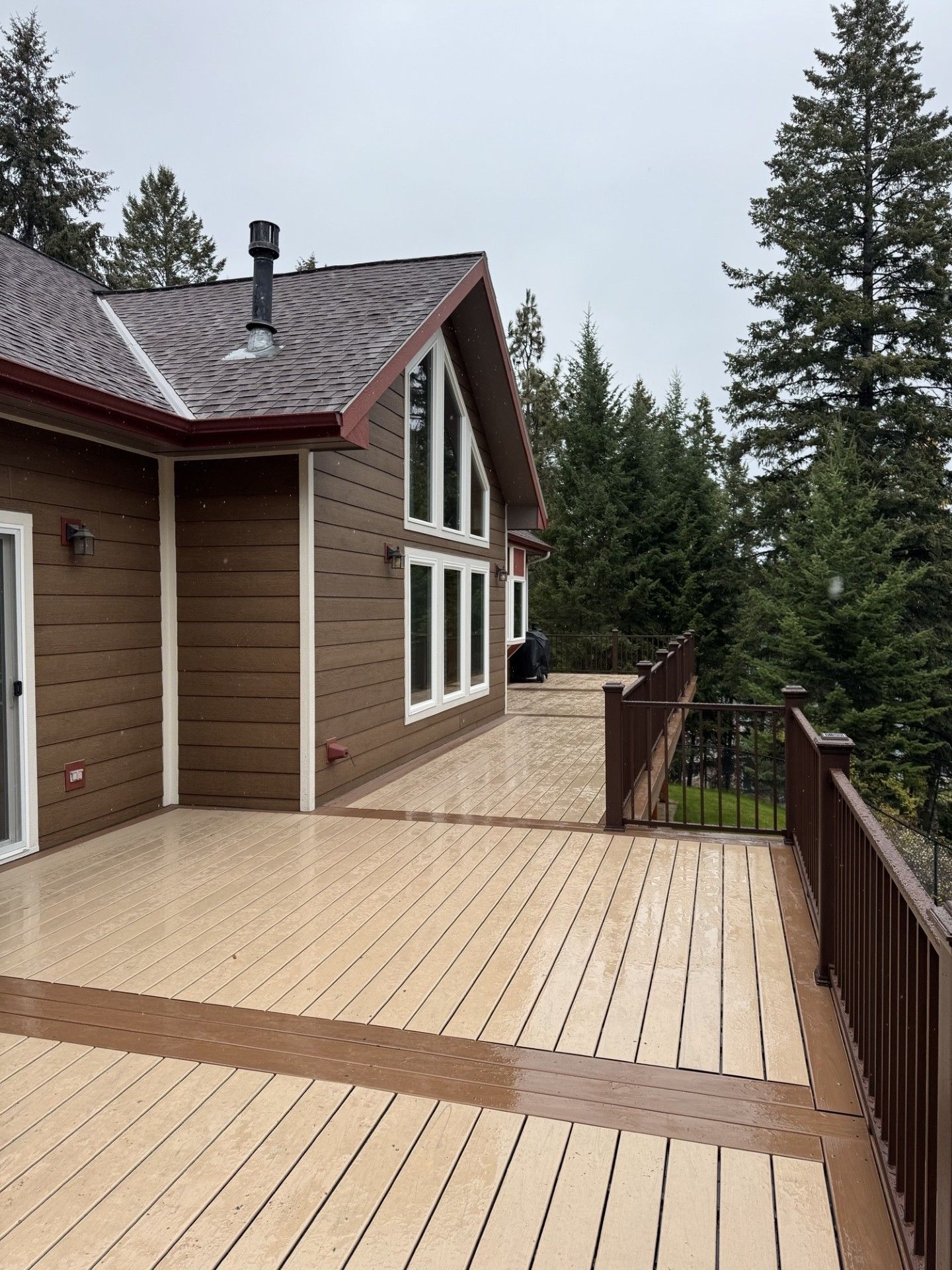 Wooden deck beside a brown house, with evergreen trees and a forested view beyond.