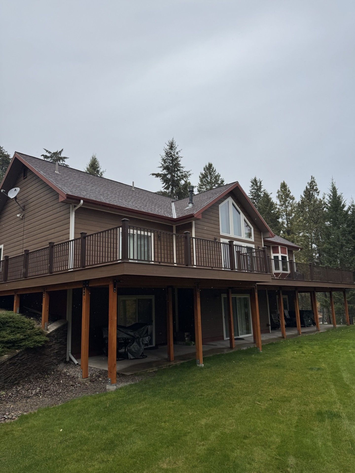 Rustic two-story house with a wraparound deck and green lawn, viewed from the side under an overcast sky