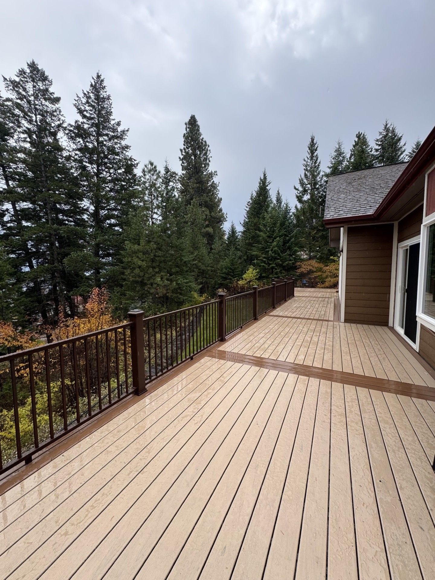 Wooden deck with railing beside a house, overlooking evergreen trees under a cloudy sky.