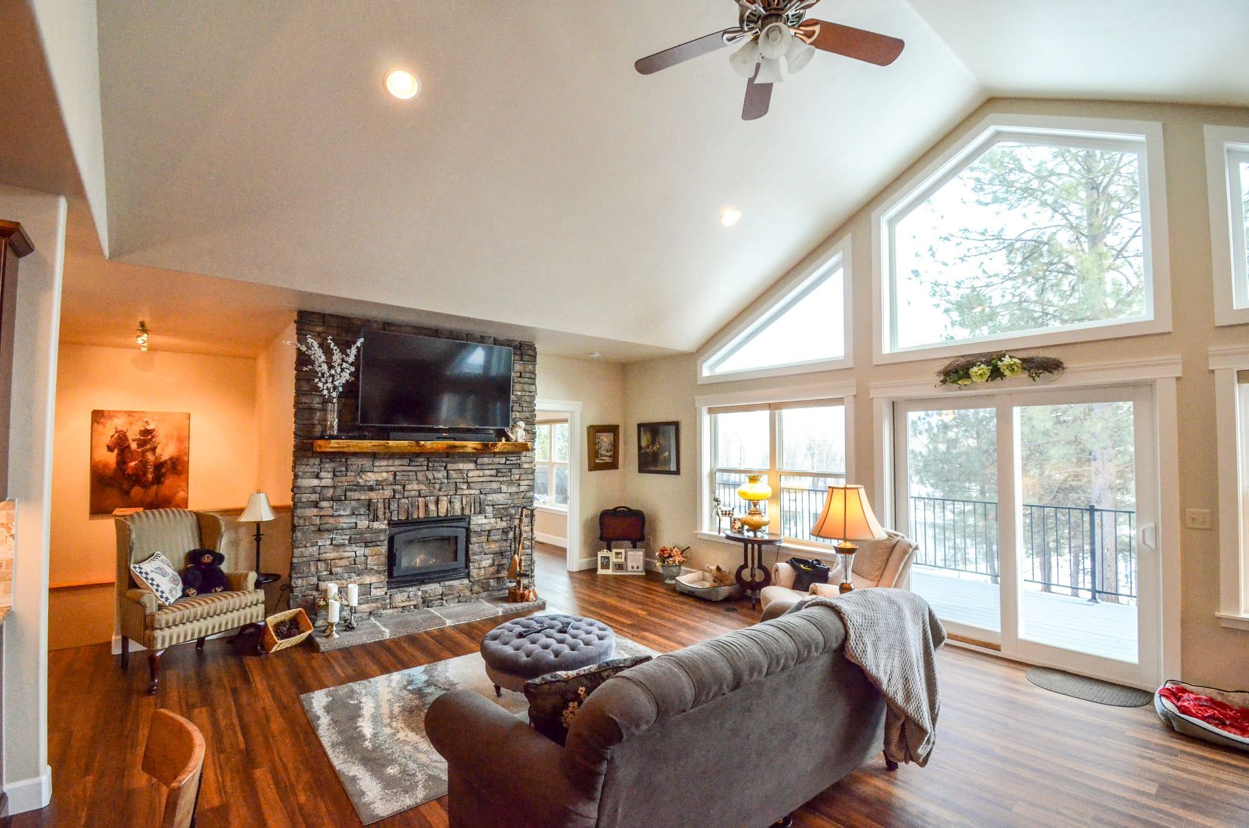 Bright living room with stone fireplace, gray sofa, hardwood floors, and tall windows overlooking a deck