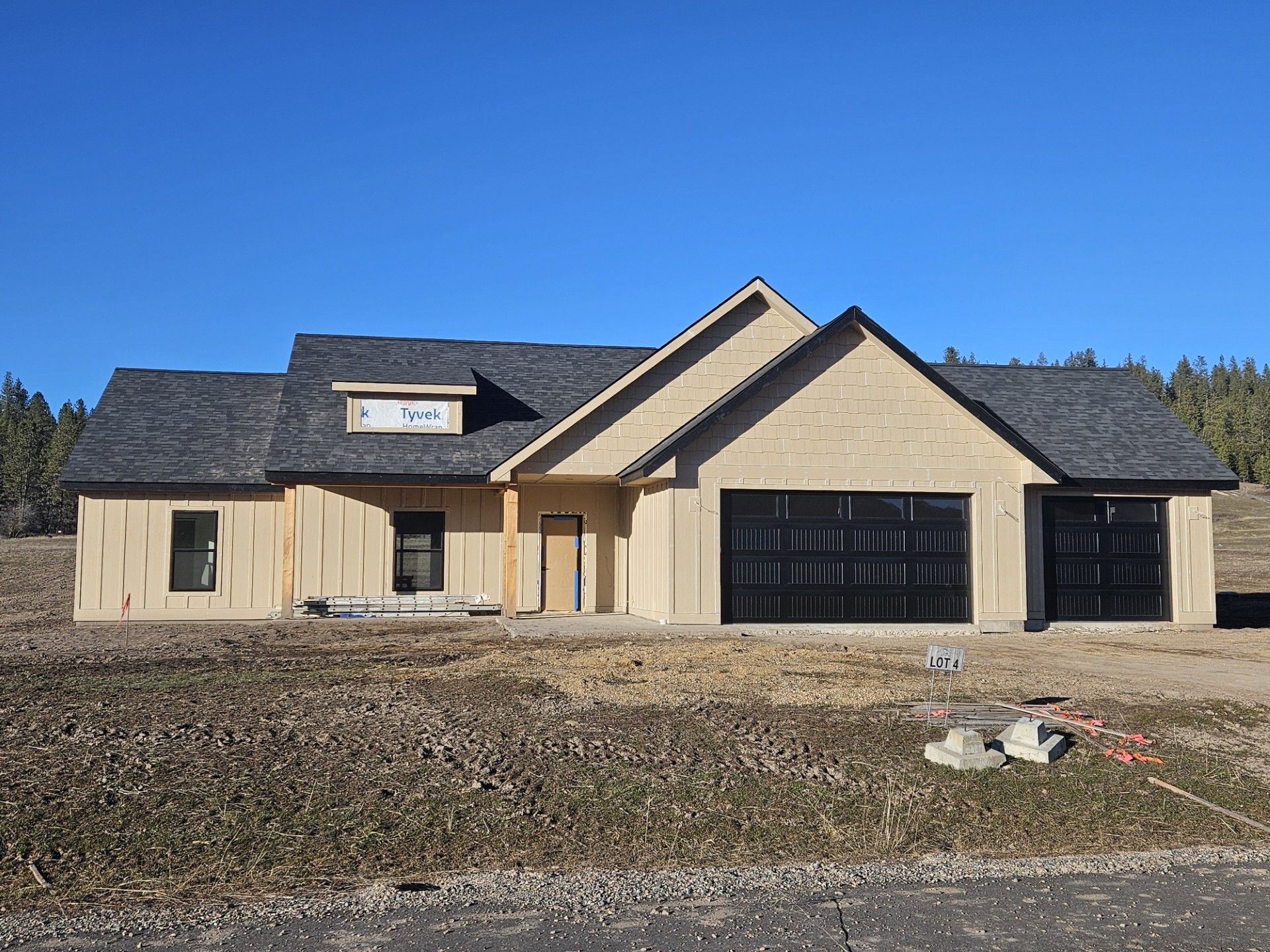 New beige house under construction with black garage doors and a large front yard under a blue sky