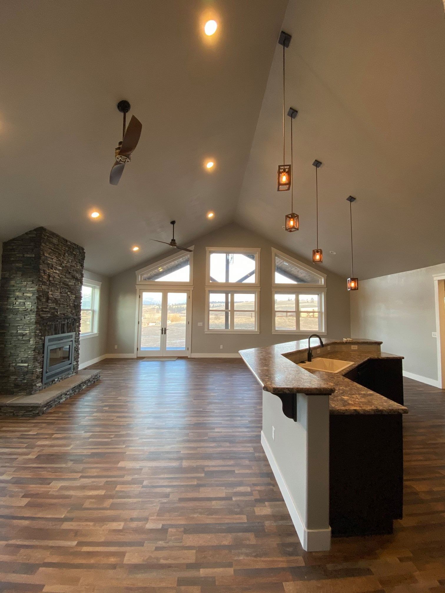 Spacious empty living room with vaulted ceiling, wood floors, stone fireplace, and large windows.