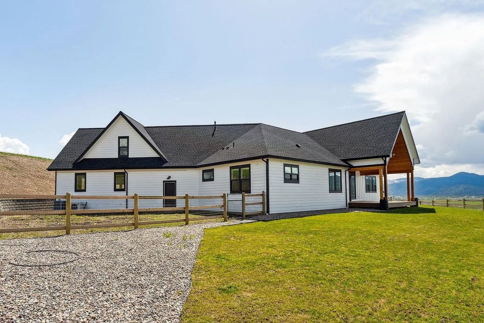 White countryside house with a dark roof, wooden fence, gravel yard, and green lawn under a blue sky