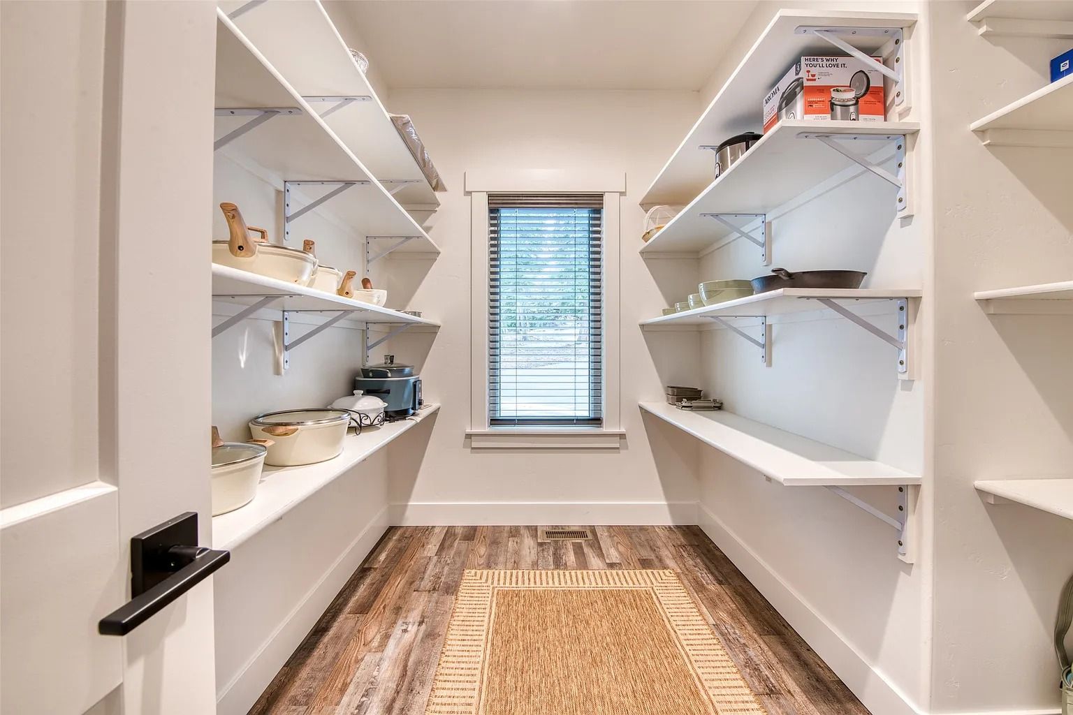 Walk-in pantry with white shelves, kitchen items, and a woven rug, lit by a small window.