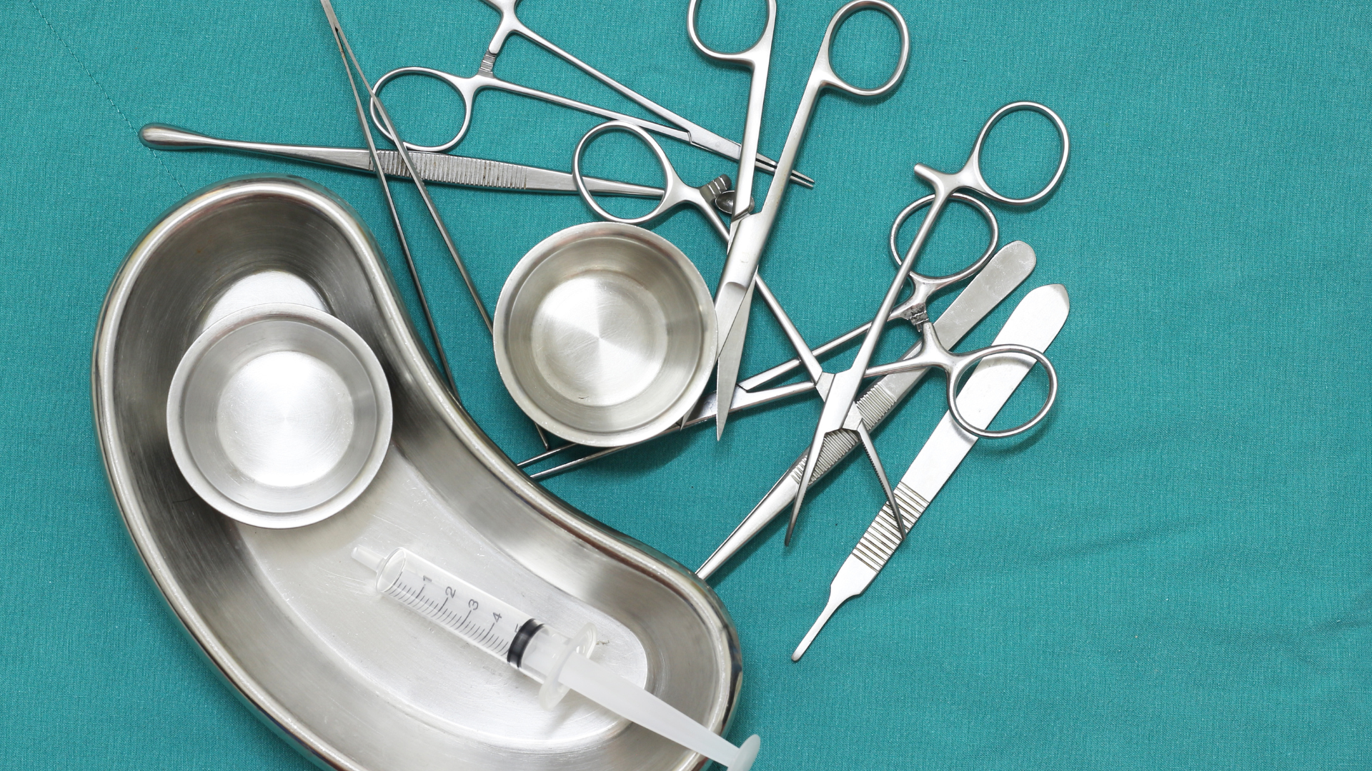 Surgical instruments on a green surface: scissors, clamps, scalpel, syringe, bowls, and a tray.