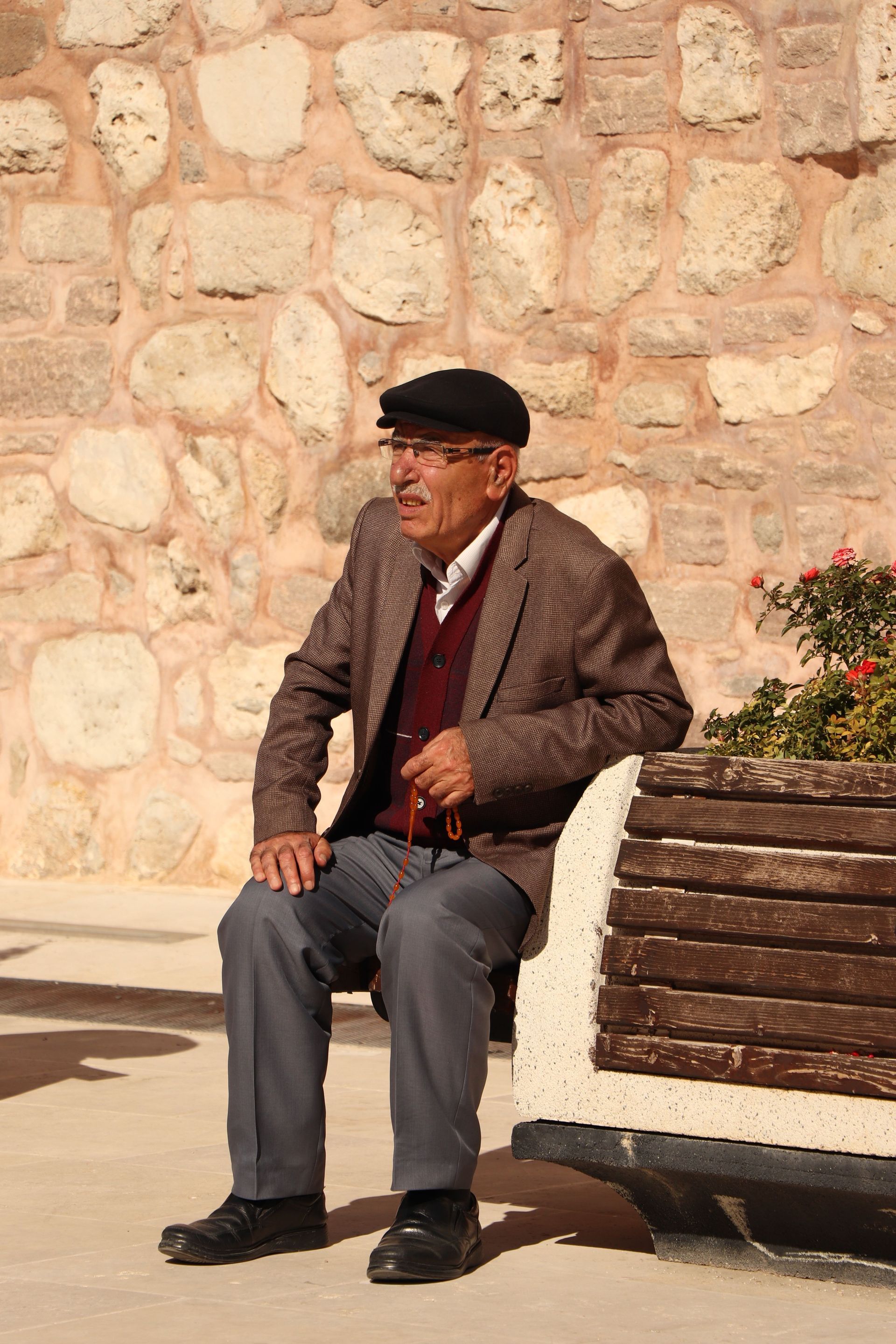 Man sitting on a bench outdoors beside a stone wall, wearing a cap and jacket.