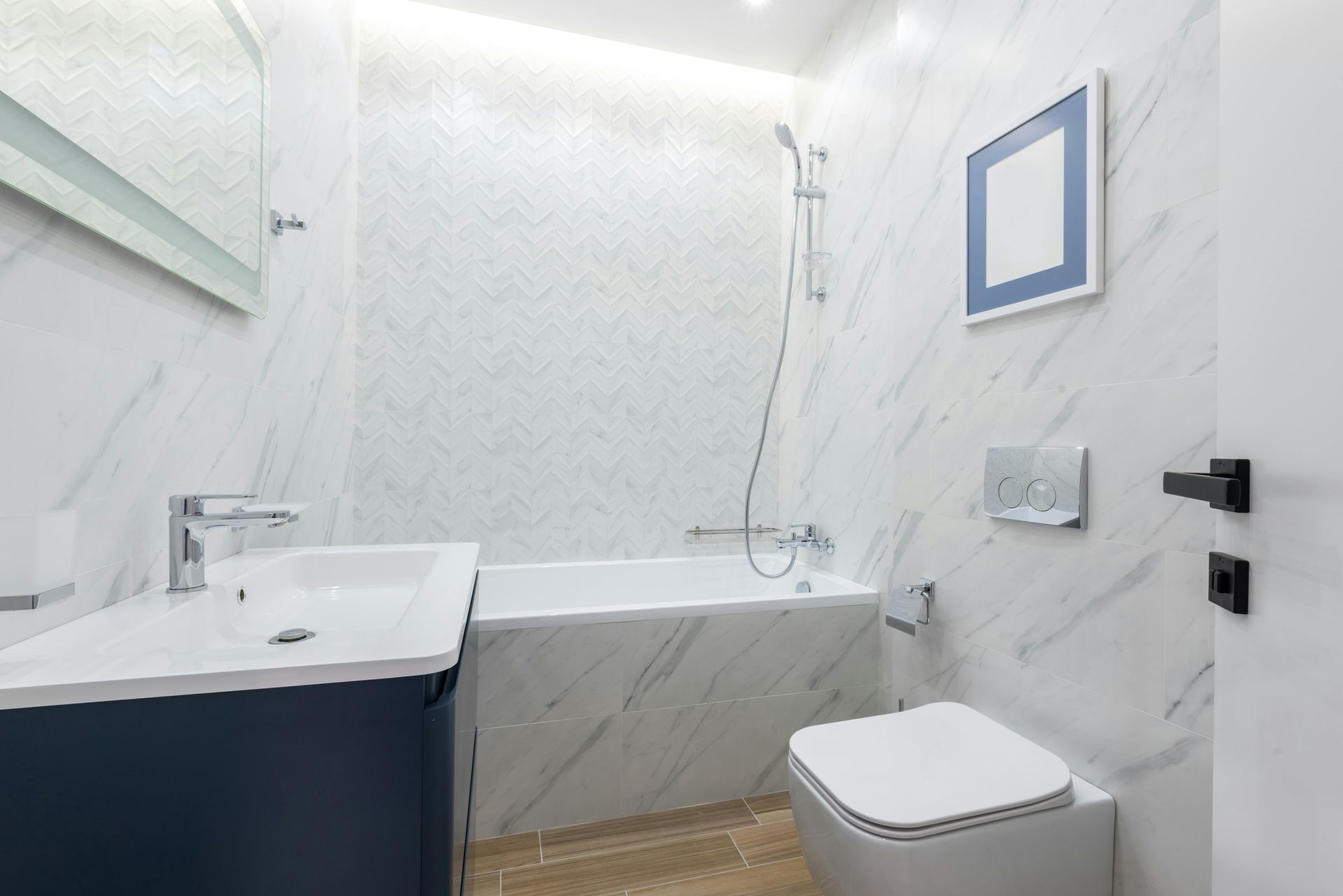 Modern bathroom with marble-tiled walls, a white vanity with a blue base, a bathtub, and a wall-mounted toilet.
