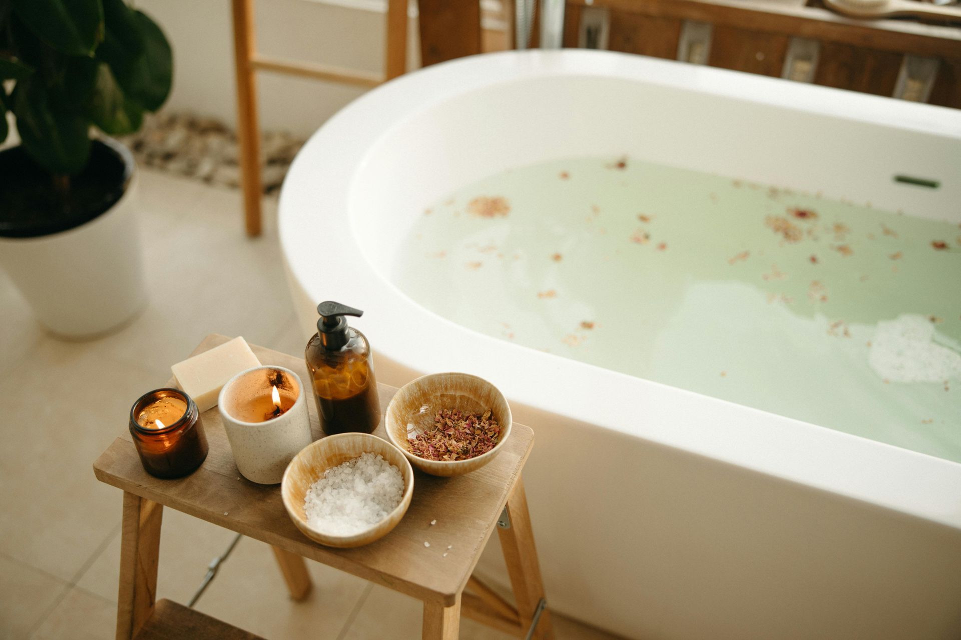 A wooden stool holds candles and bowls of bath salts next to a soaking tub filled with water and flower petals.