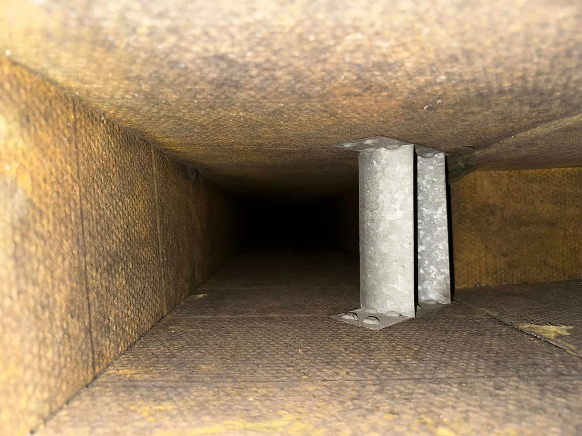 Inside view of a rectangular duct, light brown insulation, with a silver metal support in the center. Dark tunnel ahead.