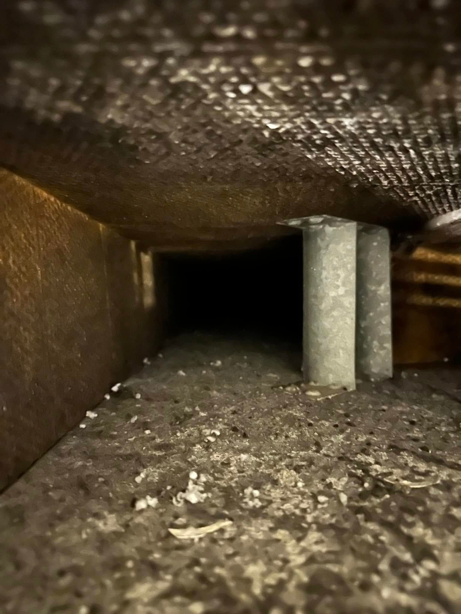 Inside a dirty air duct, dust and debris cover the bottom. Two gray, cylindrical supports are visible.