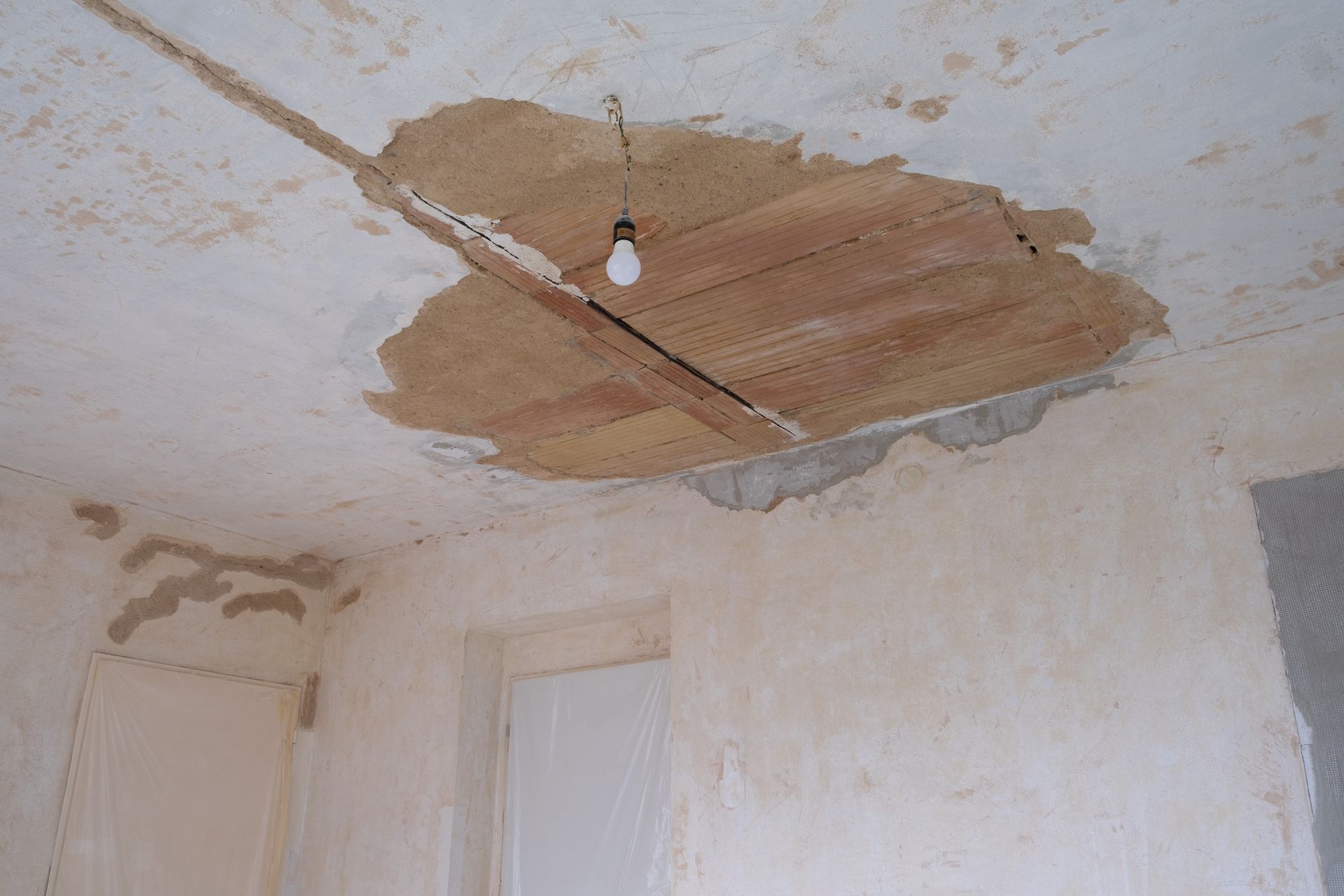 Damaged ceiling with exposed wooden beams, a lightbulb hangs, peeling walls.