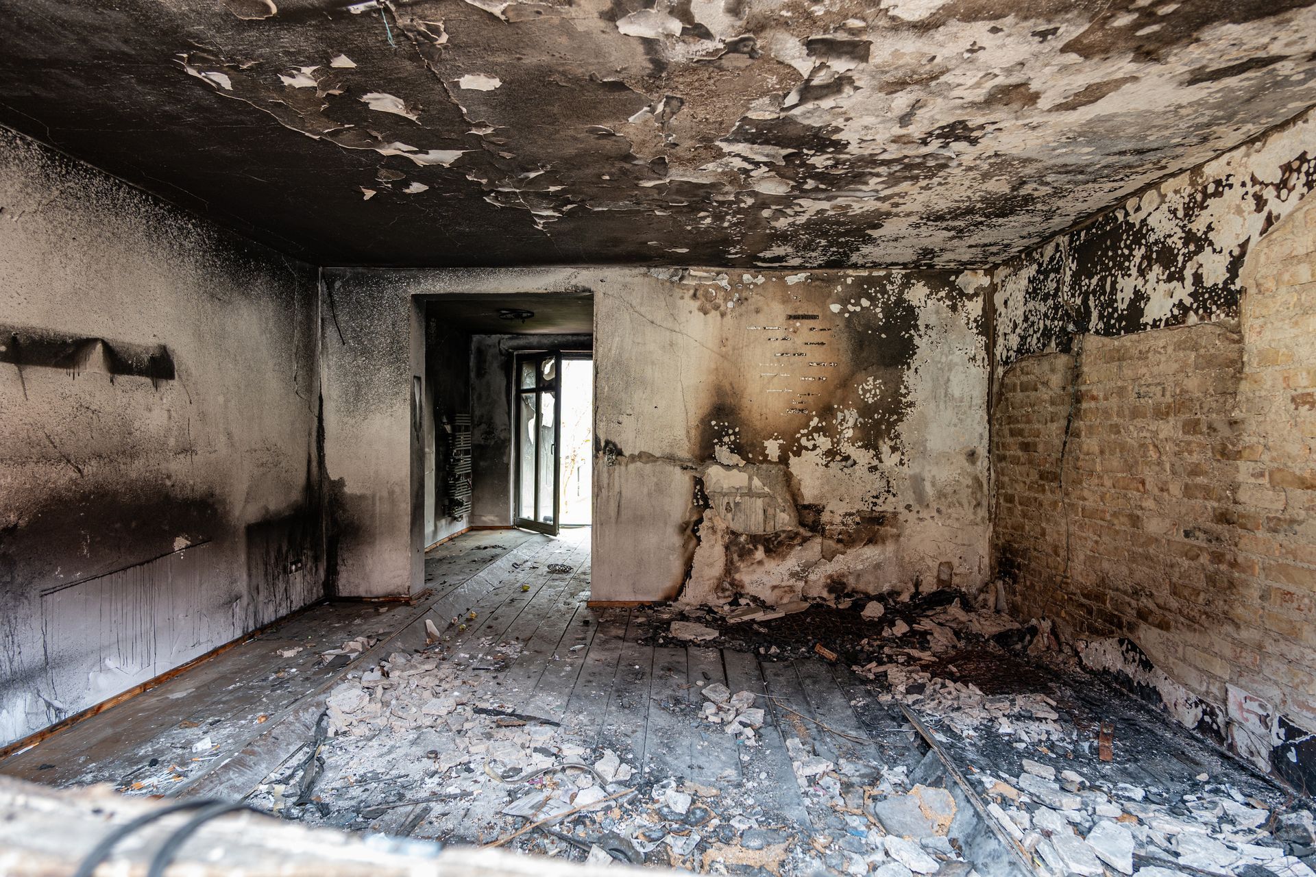 Interior of a fire-damaged room, with charred walls, peeling paint, and a doorway leading outside.