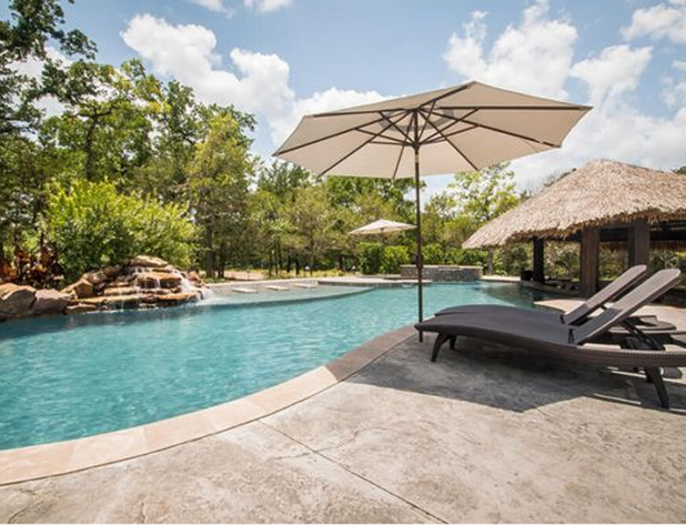 Swimming pool with waterfall, sun umbrella, lounge chairs, and tiki hut. Lush green trees in background.