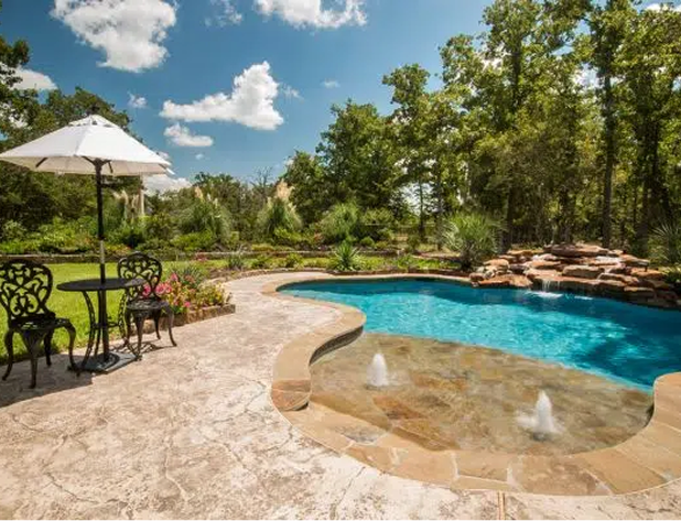A backyard pool with a rock waterfall, patio table and umbrella, on a sunny day.