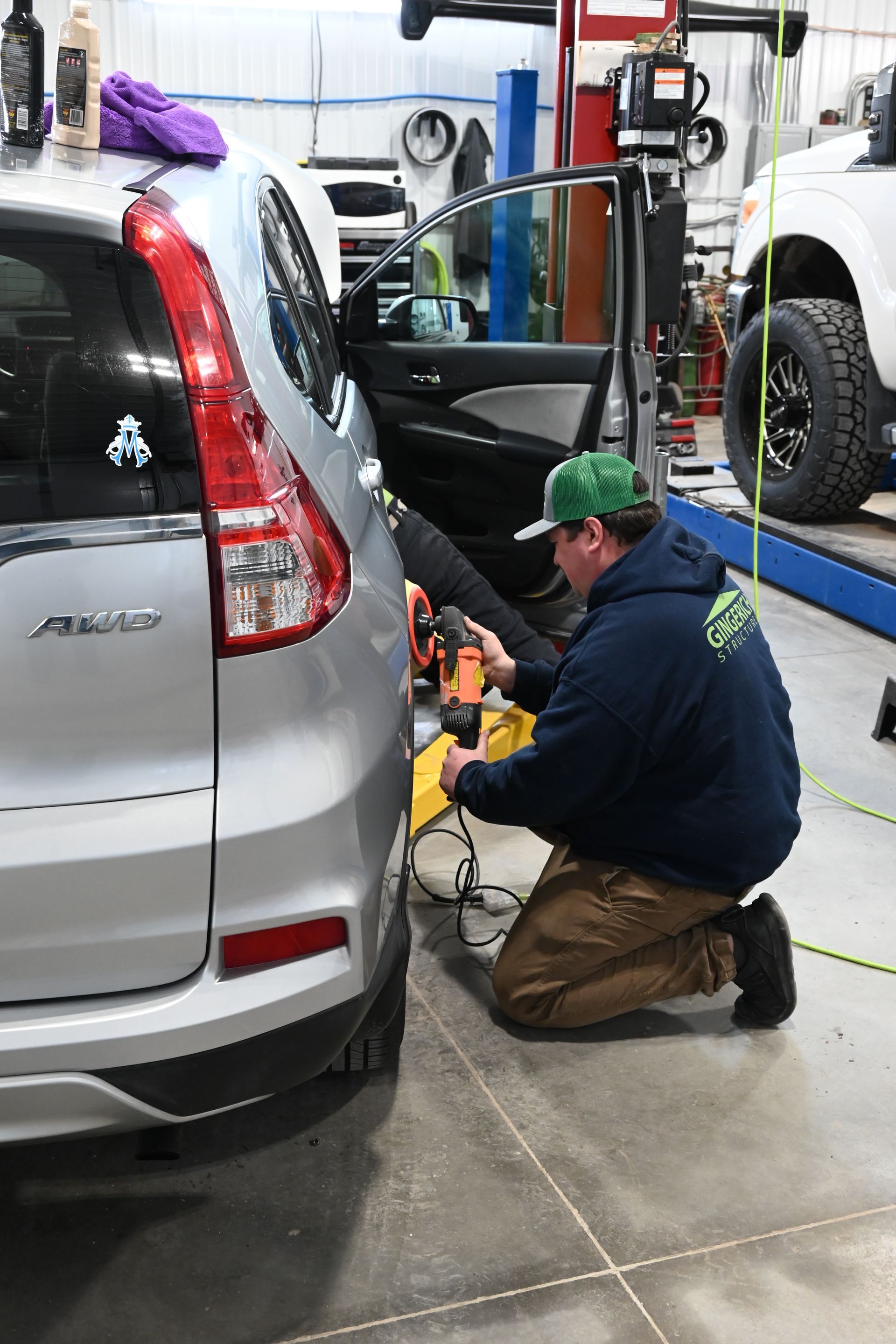 A person in a green cap and navy hoodie kneels to buff the side of a silver SUV inside an auto repair garage.