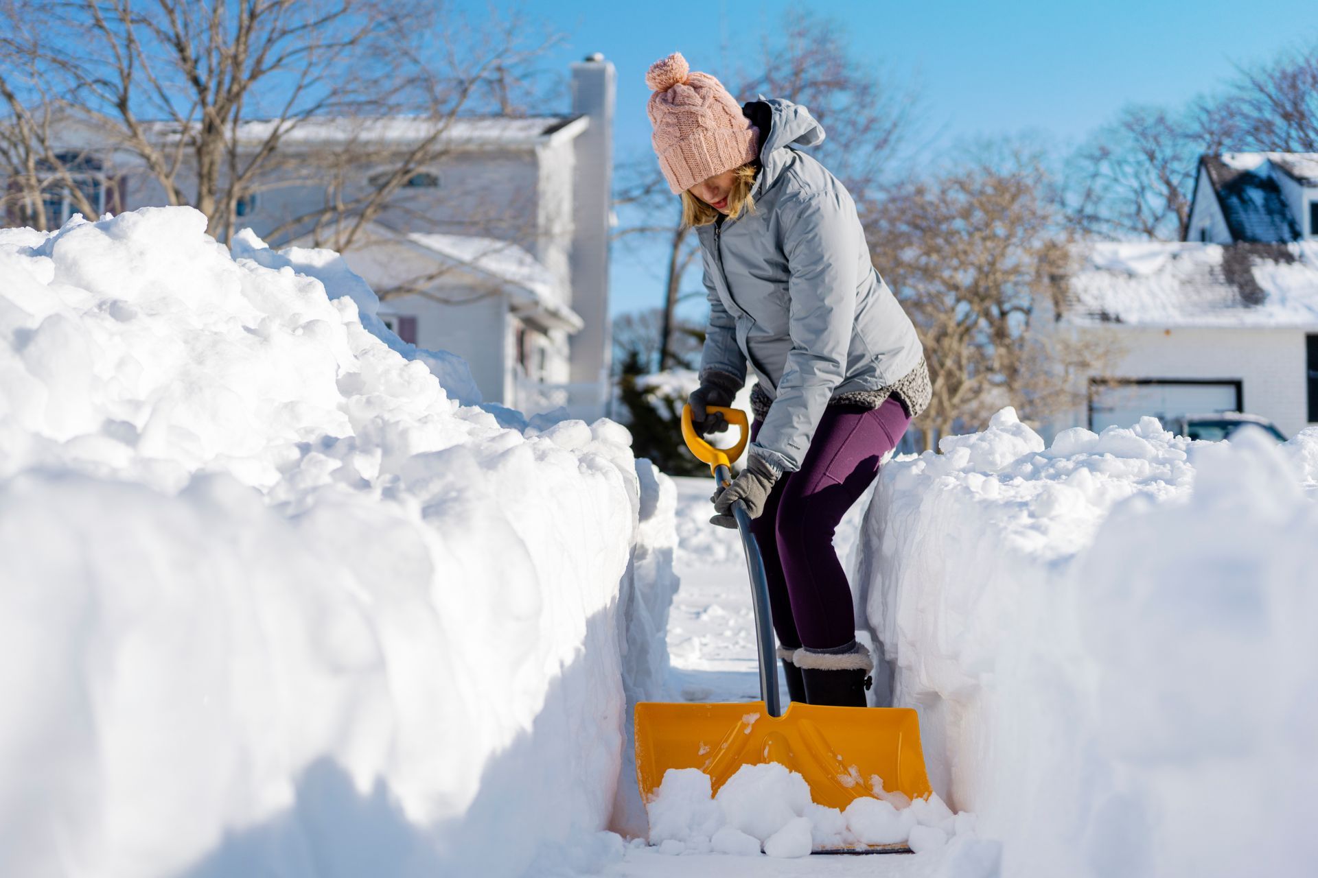Person shoveling snow on a sidewalk between tall snowbanks, wearing a pink hat and gray jacket.