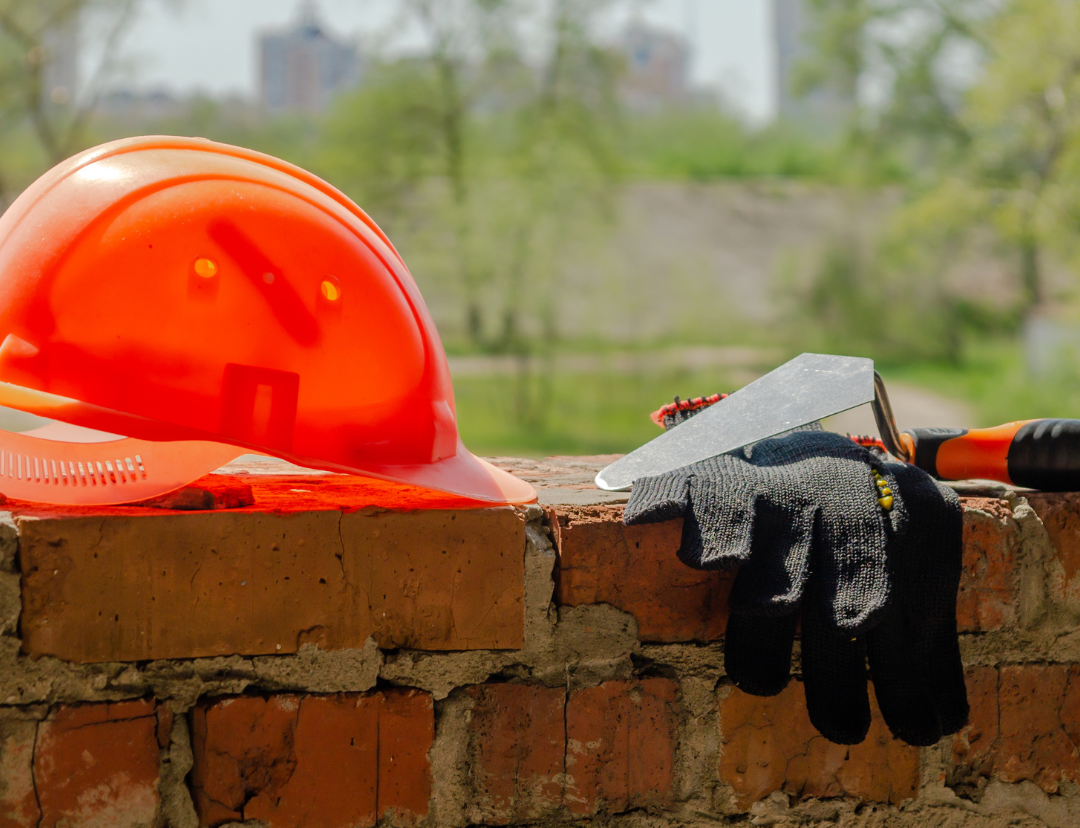 Casque de sécurité orange, gants de travail et truelle sur un mur de briques avec un fond vert flou.