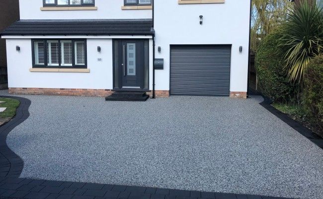 A white house with a black garage door and a grey resin driveway in front of it. Installed by Essex Resin driveways