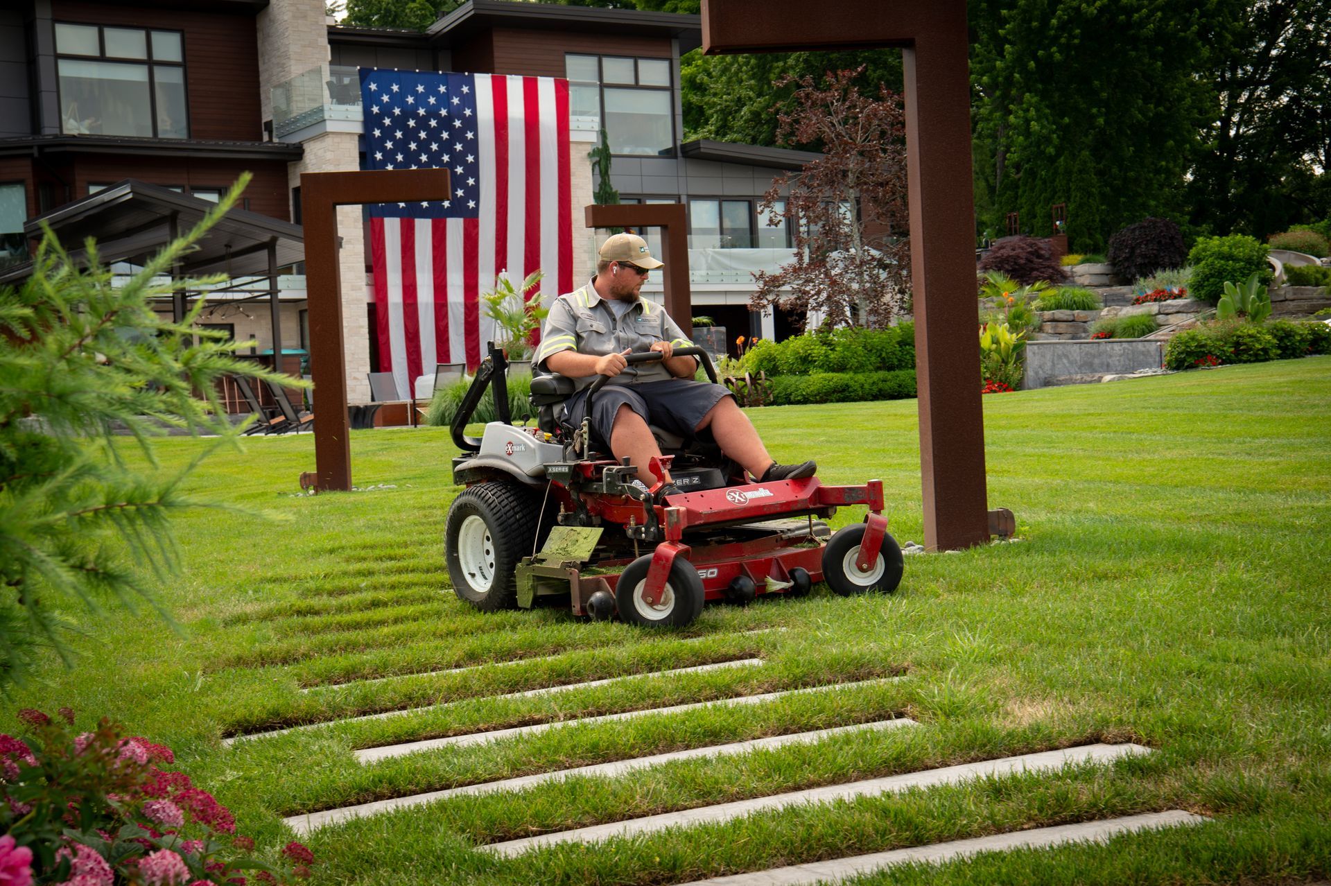 Man on riding lawnmower cutting grass in front of a house with American flag.