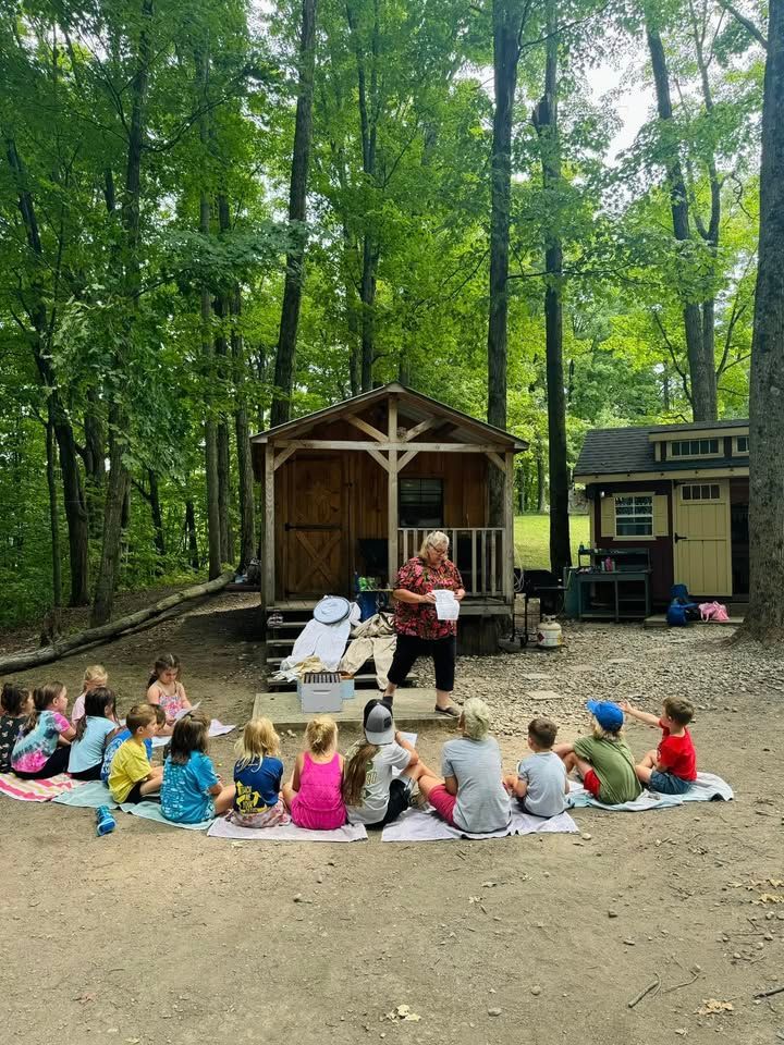 Group of children listening to a woman reading outside, near a cabin in a wooded area.