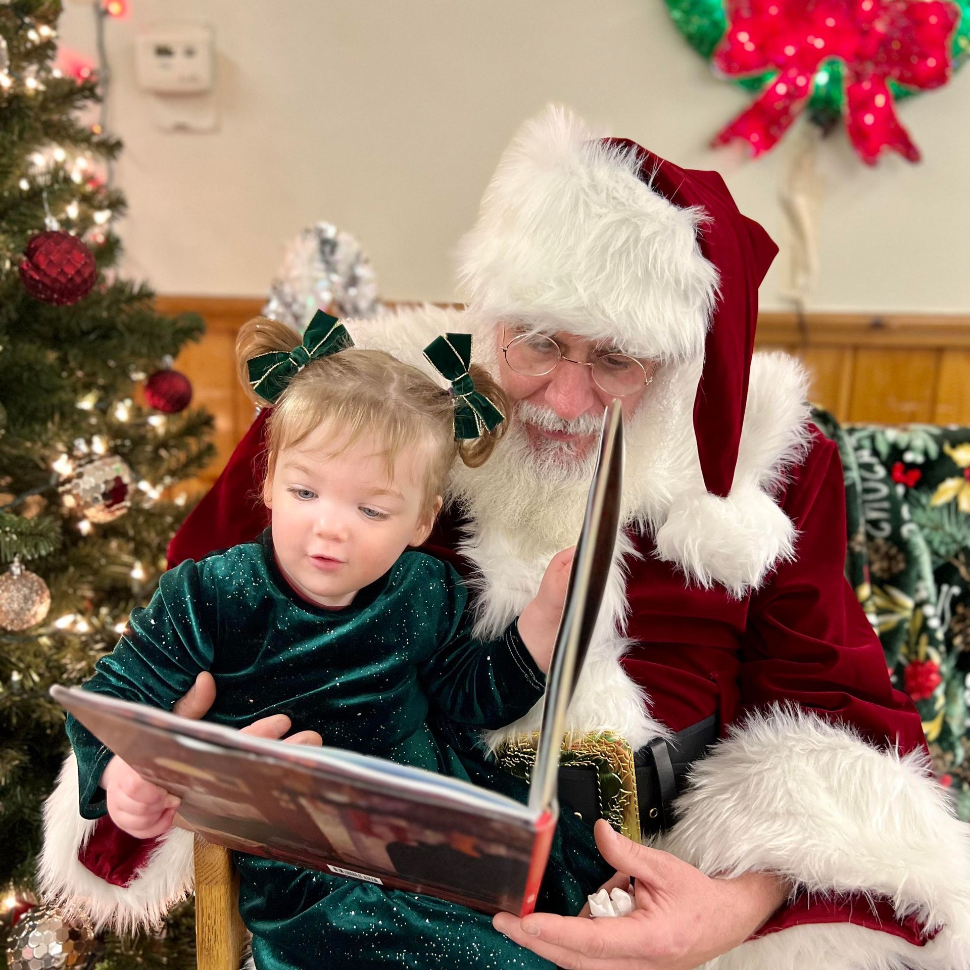 Santa Claus reads a book with a young girl, both focused, by a Christmas tree.