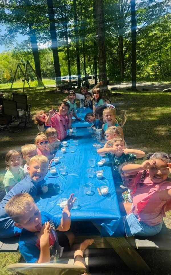 Children at a long picnic table outdoors, smiling and posing. Blue tablecloth, sunny day, surrounded by trees.