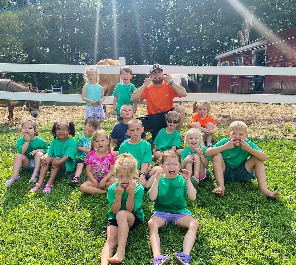 A group of children and an adult making silly faces, sitting on grass in front of a white fence with animals.