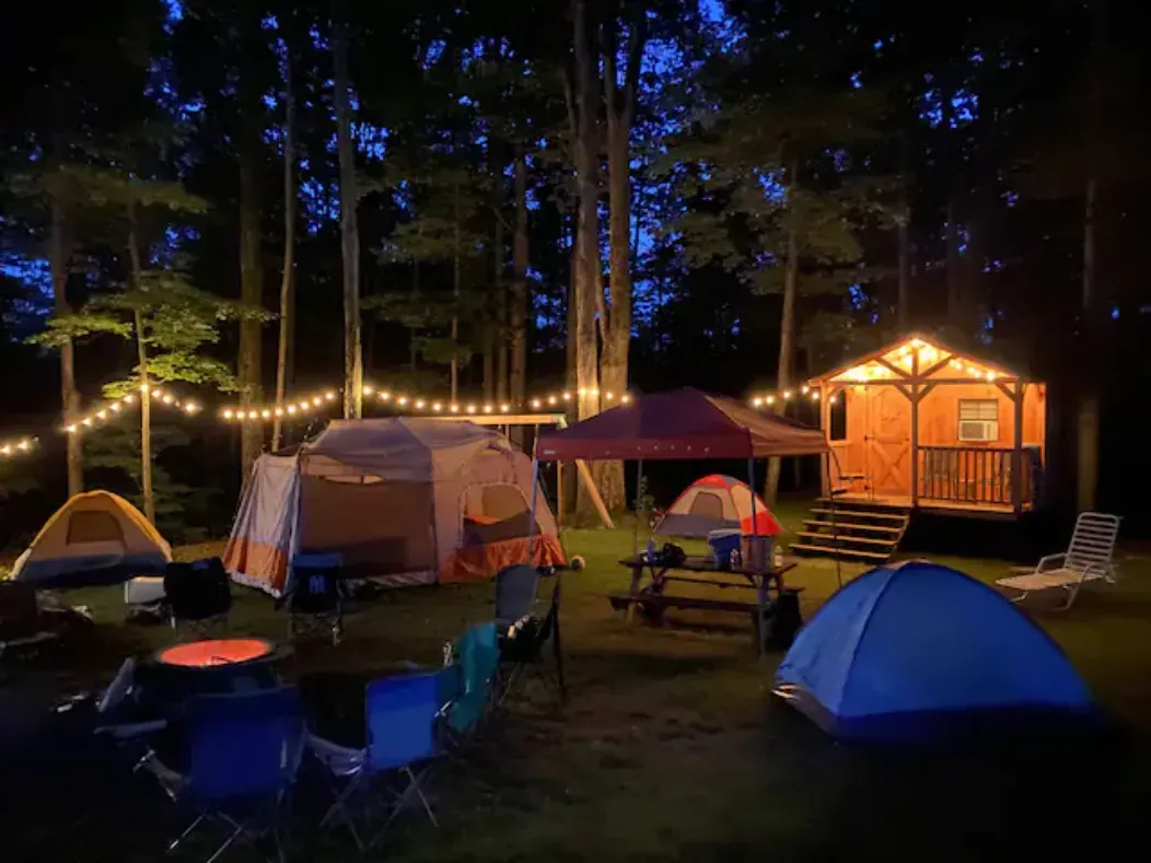 Camping scene at night with tents, a cabin, and string lights in a wooded area.
