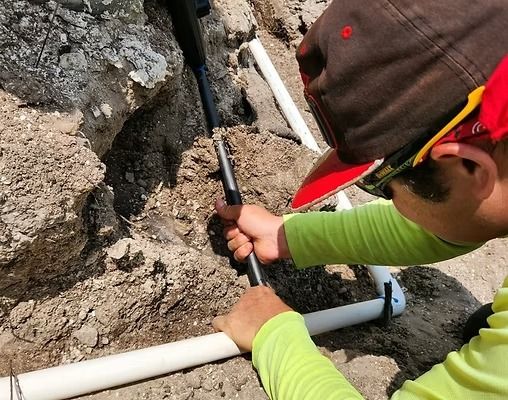 Person in a cap and sunglasses using a tool on buried pipes in a trench.