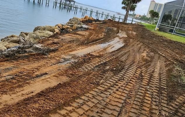Shoreline construction site with mud, rocks, and machine tracks near a body of water and pier.
