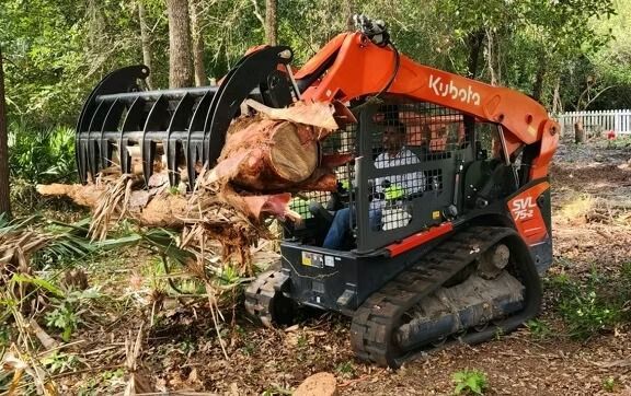 Orange Kubota skid steer with grapple bucket, picking up logs in a wooded area.