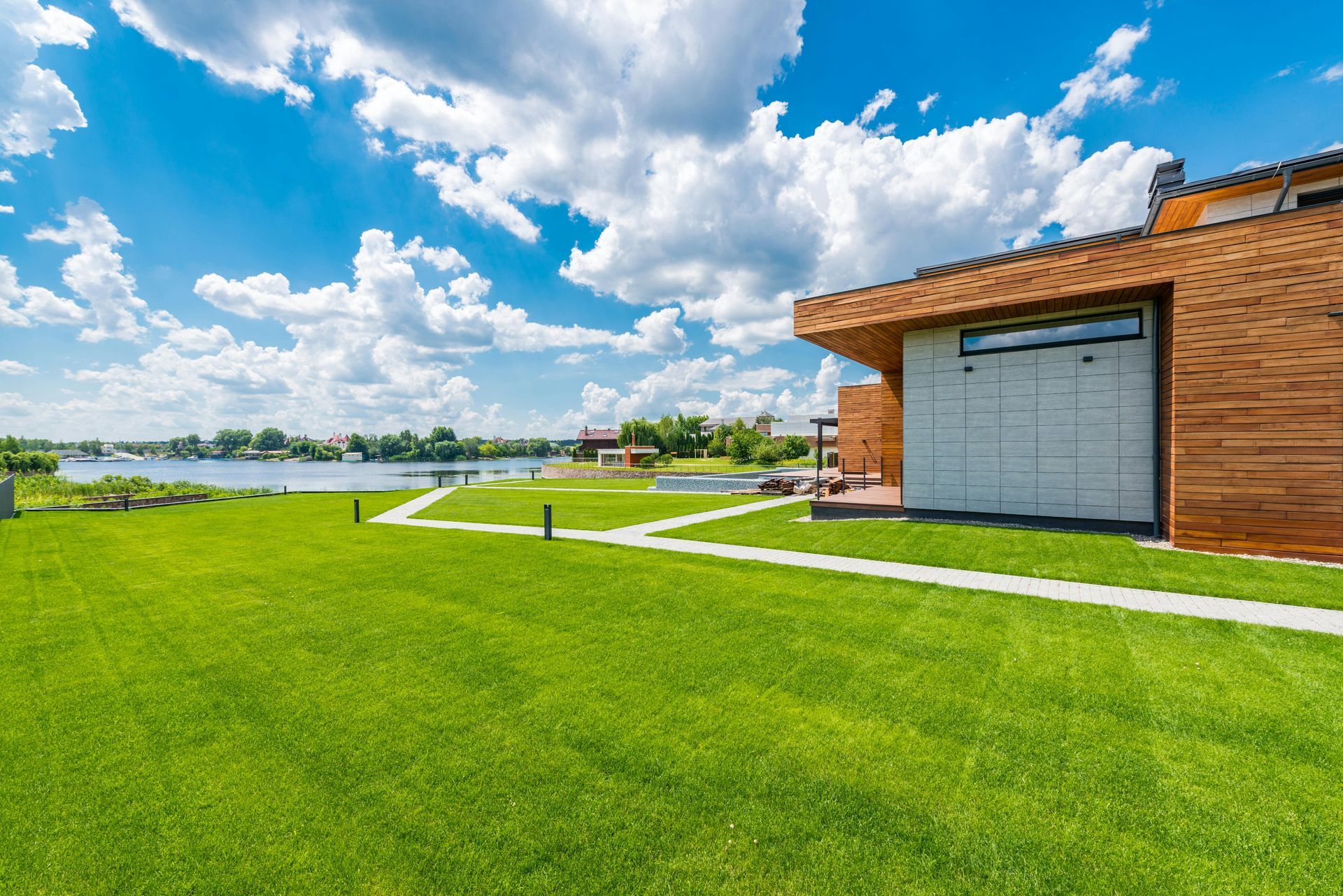 A modern house with wooden siding stands beside a vibrant green lawn under a blue, cloudy sky, overlooking a wide river.