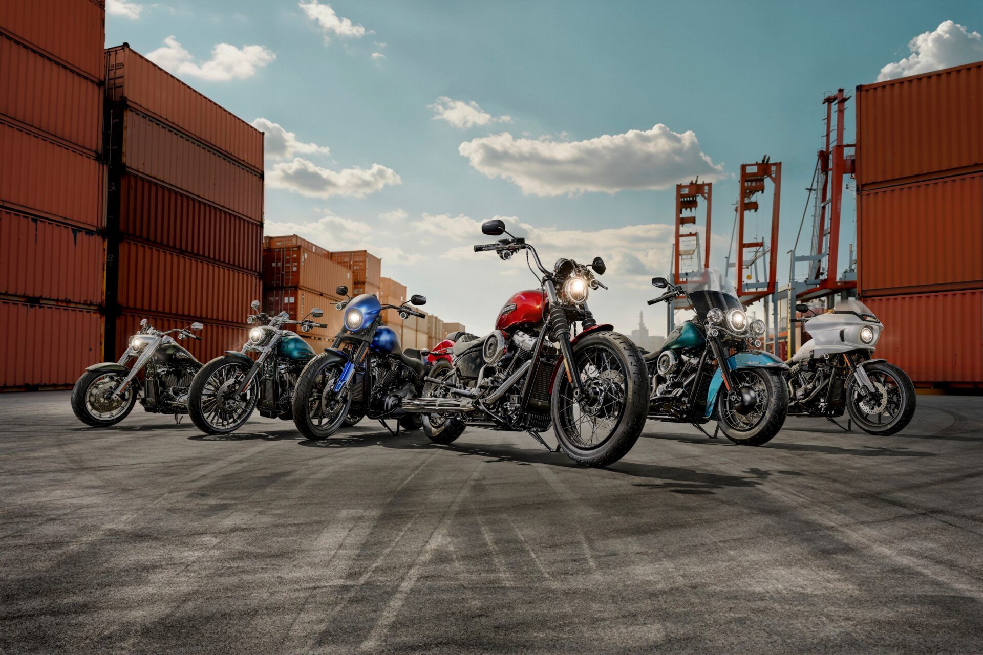 Cinco motocicletas enfileiradas em uma doca ao lado de contêineres empilhados sob um céu parcialmente nublado.