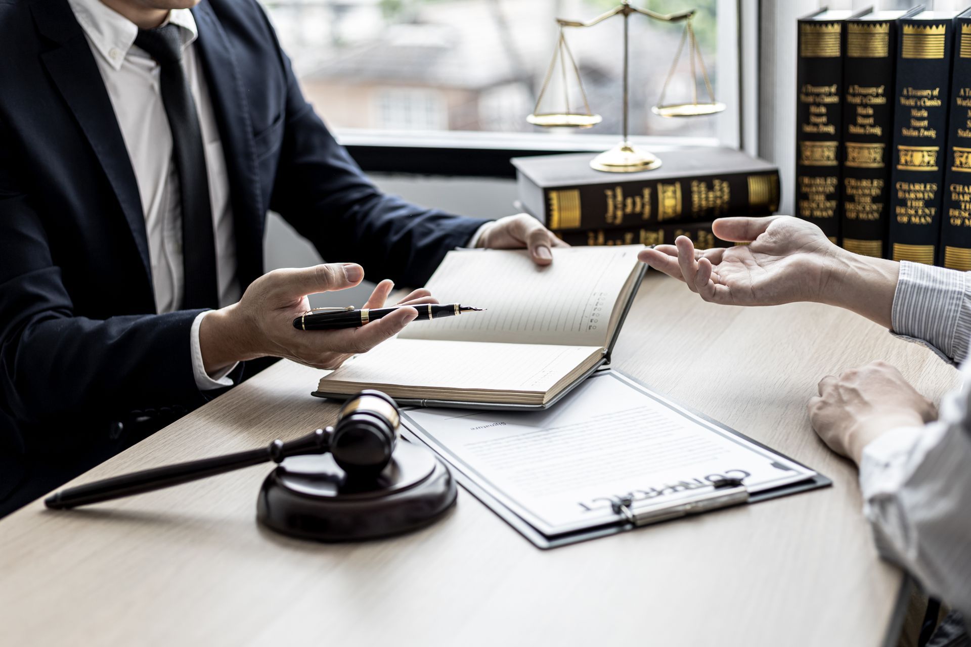 A legal consultation between two people at a desk with law books, a gavel, and scales of justice.