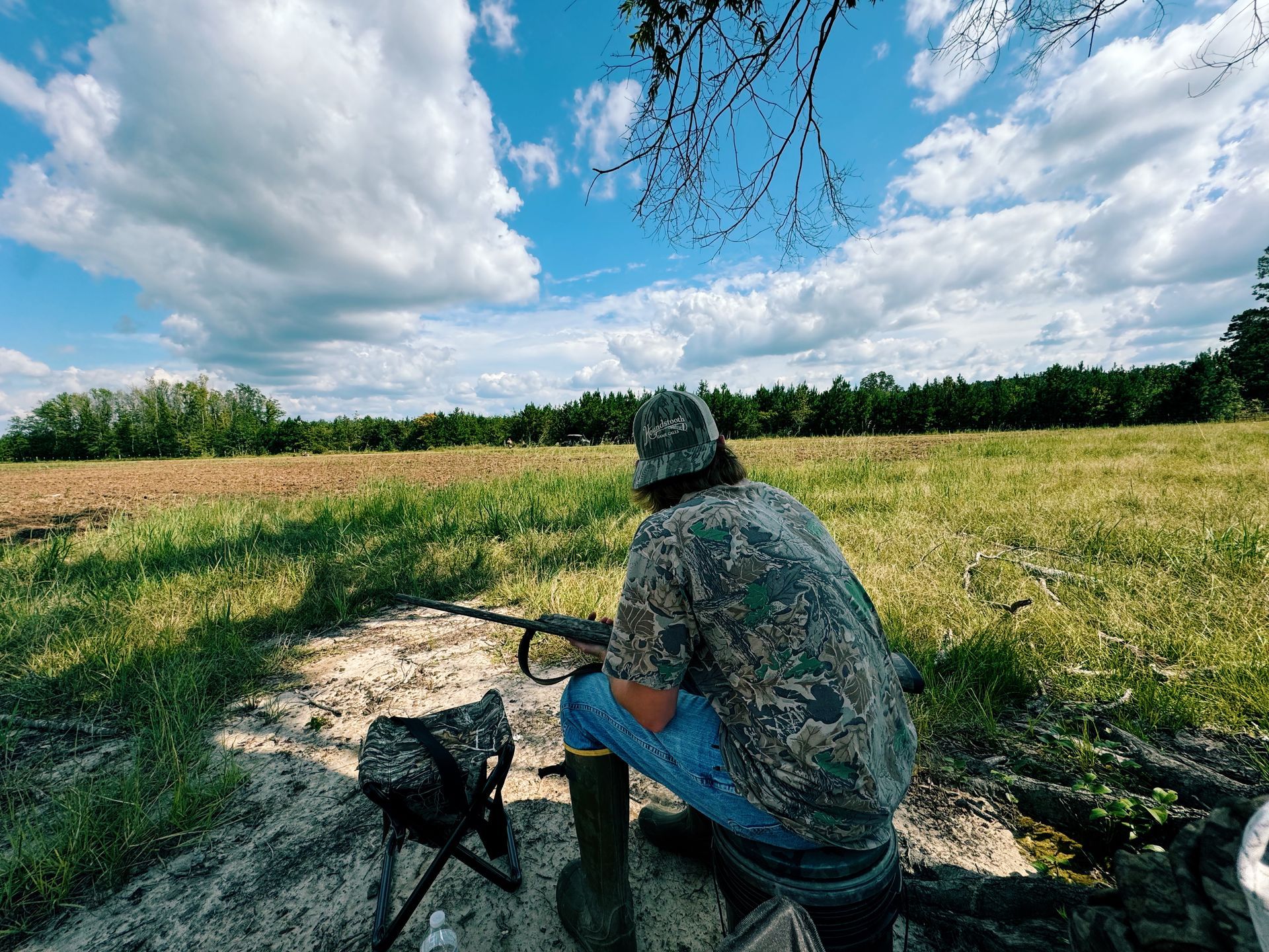 Hunter kneeling in field, aiming shotgun; camouflage clothing, bright sky, field.